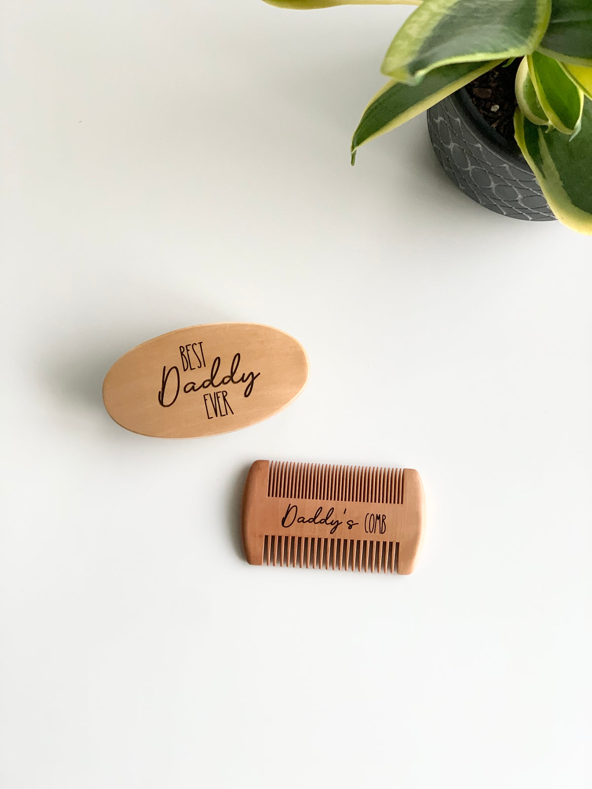 Custom wooden beard brush engraved with Best Daddy Ever and wooden comb engraved Daddy’s Comb on white surface near green plant pot