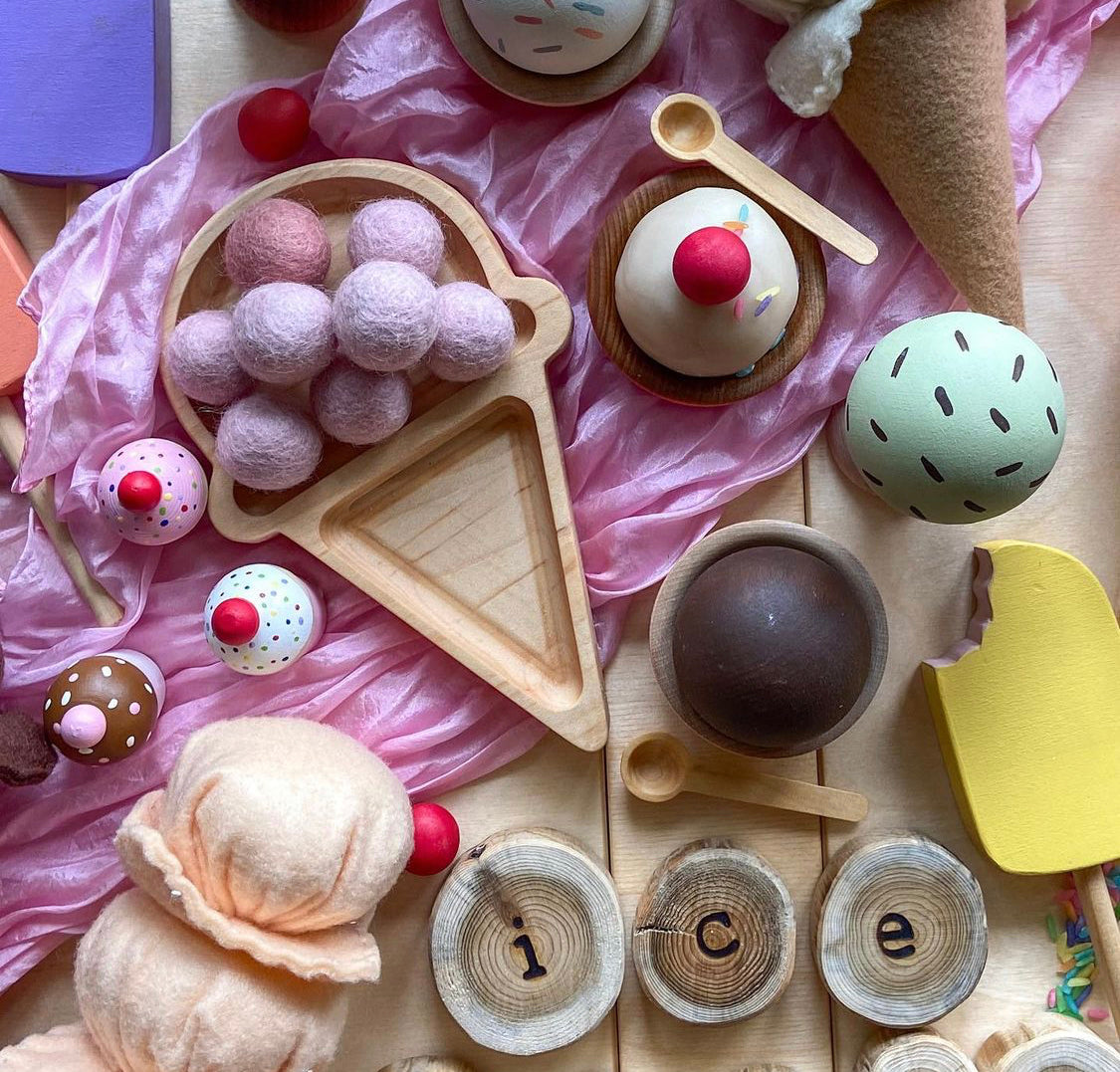 Wooden ice cream shaped sensory tray with felt ice cream scoops and assorted wooden dessert toys on pink fabric background
