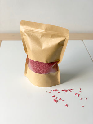Brown kraft resealable pouch filled with pink coloured sensory rice with some grains spilled on white surface