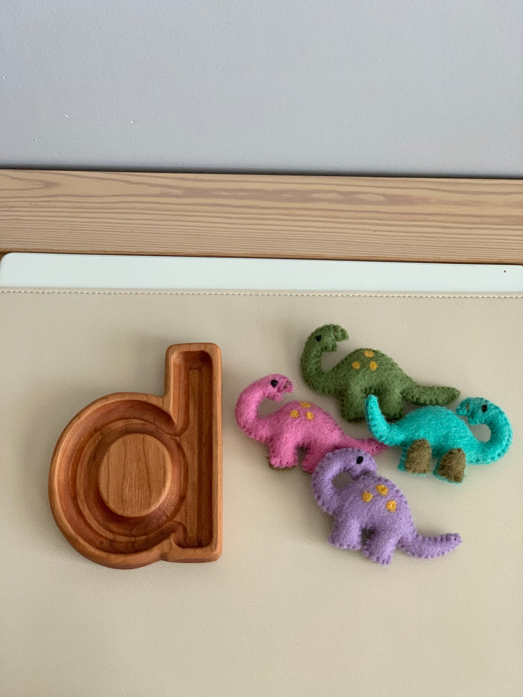 Natural wood letter d sensory tray with four small colorful felt dinosaur toys on beige surface
