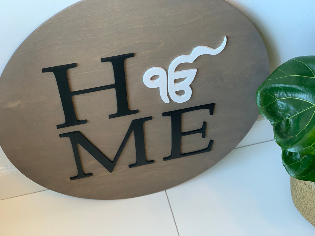 Oval custom 3D wooden name sign with black and white lettering spelling HOME on light brown wood background with green plant nearby