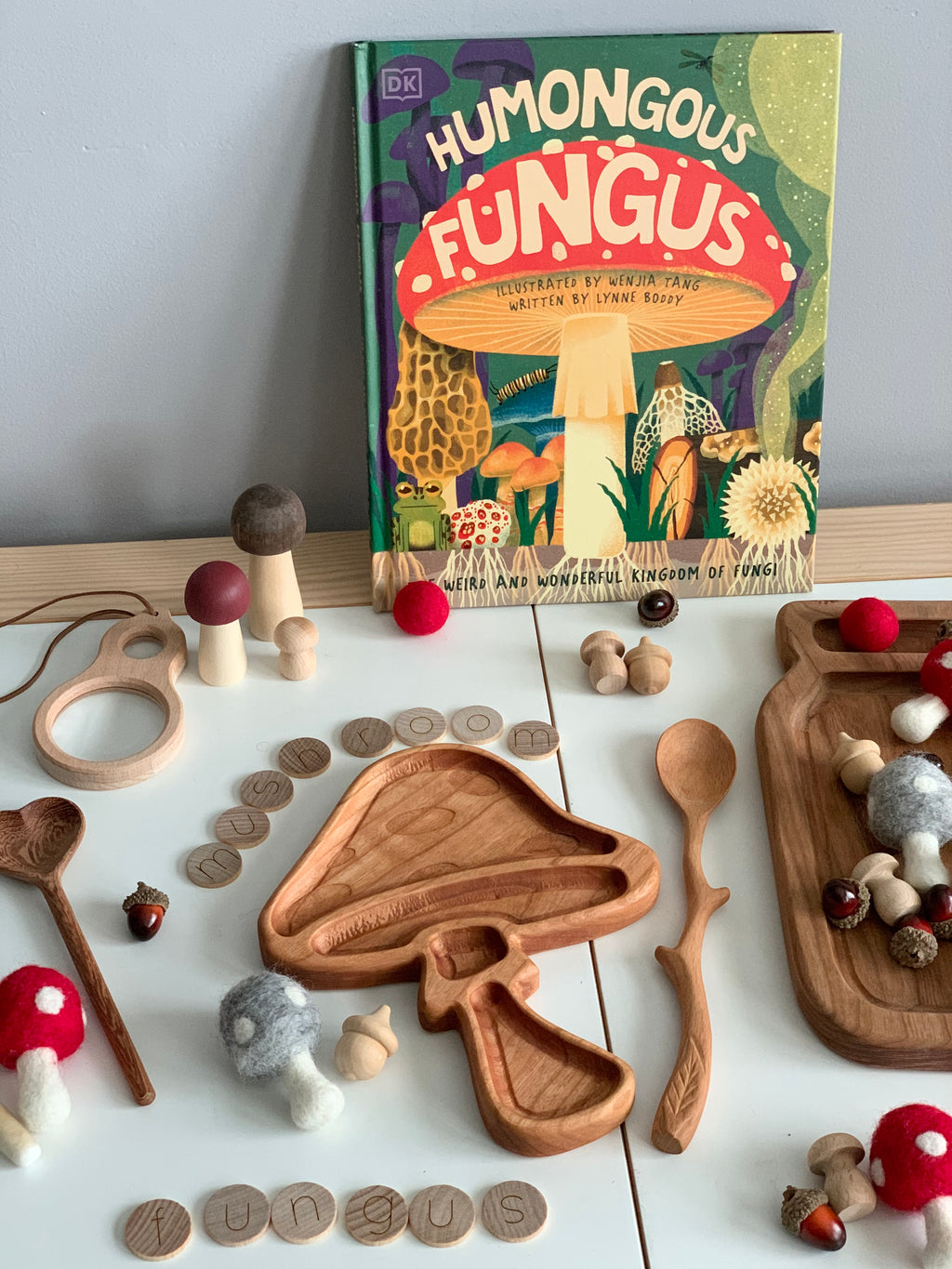 Wooden mushroom-shaped sensory tray with wooden letters, spoons, felt balls, small wooden mushrooms, and fungi book on white table