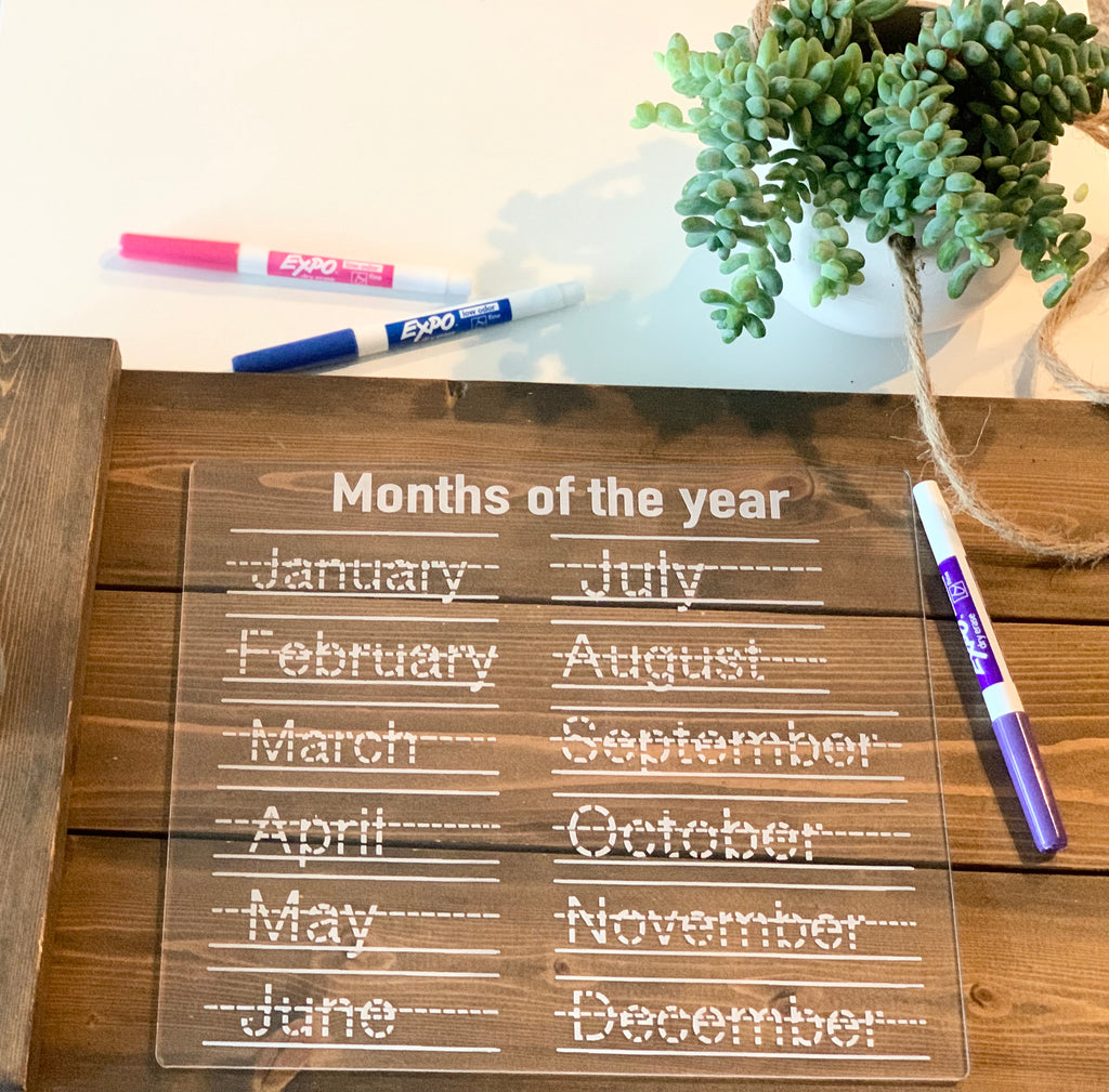 Clear acrylic dry erase board with months of the year in dotted tracing font on wooden surface with markers and plant