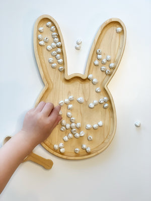 Light wooden bunny-shaped sensory tray with small white pebbles and child’s hand arranging pieces on white surface