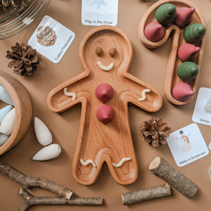 Wooden gingerbread-shaped sensory tray with painted red and white details surrounded by pinecones and nature cards