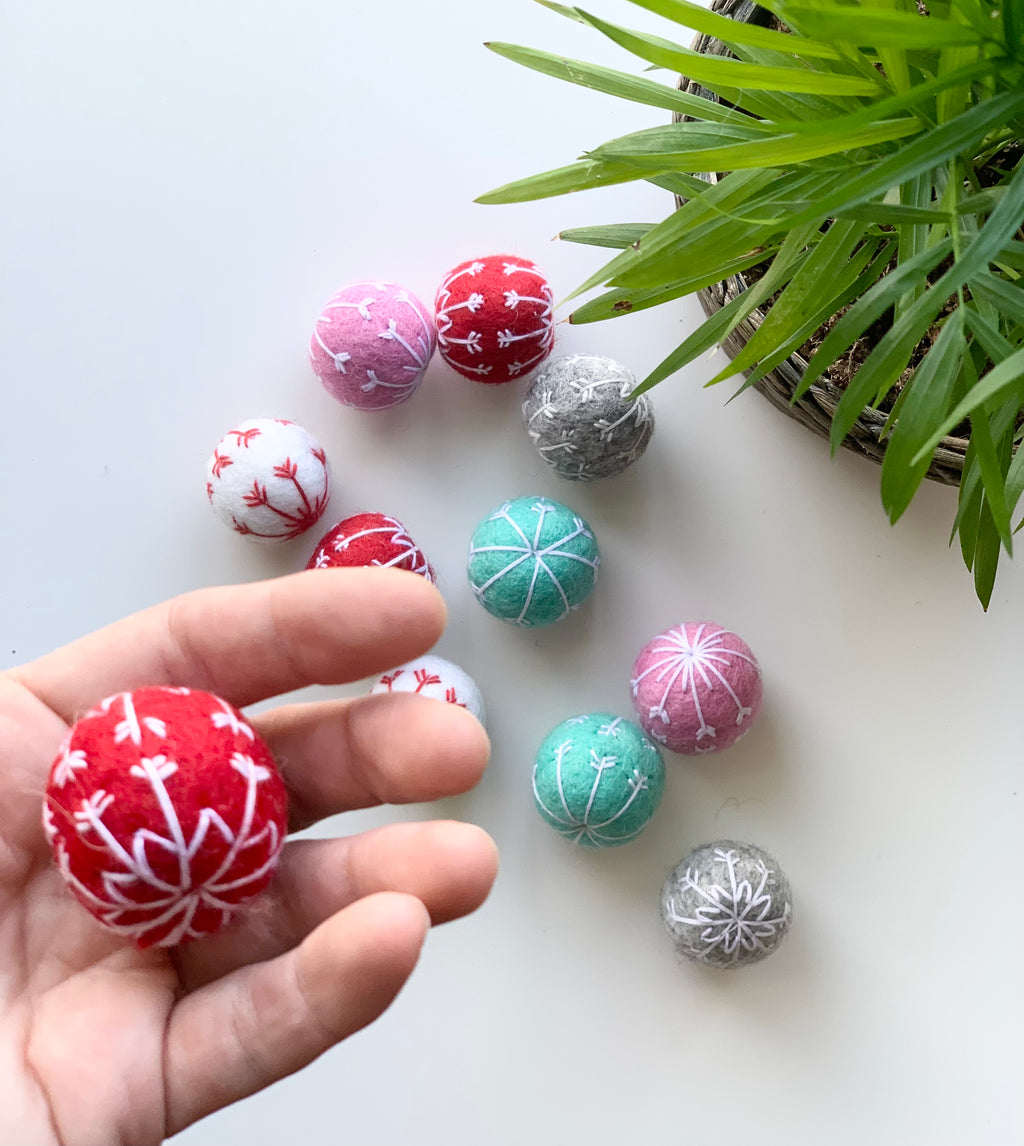 Hand holding red felt snowflake ball with white embroidery among assorted colored felt balls on white surface near green plant