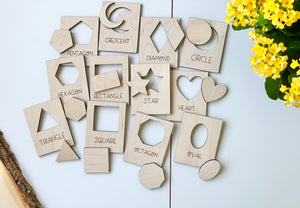 Natural wooden shape puzzle flash cards with cutouts and shape names arranged on white surface near yellow flowers