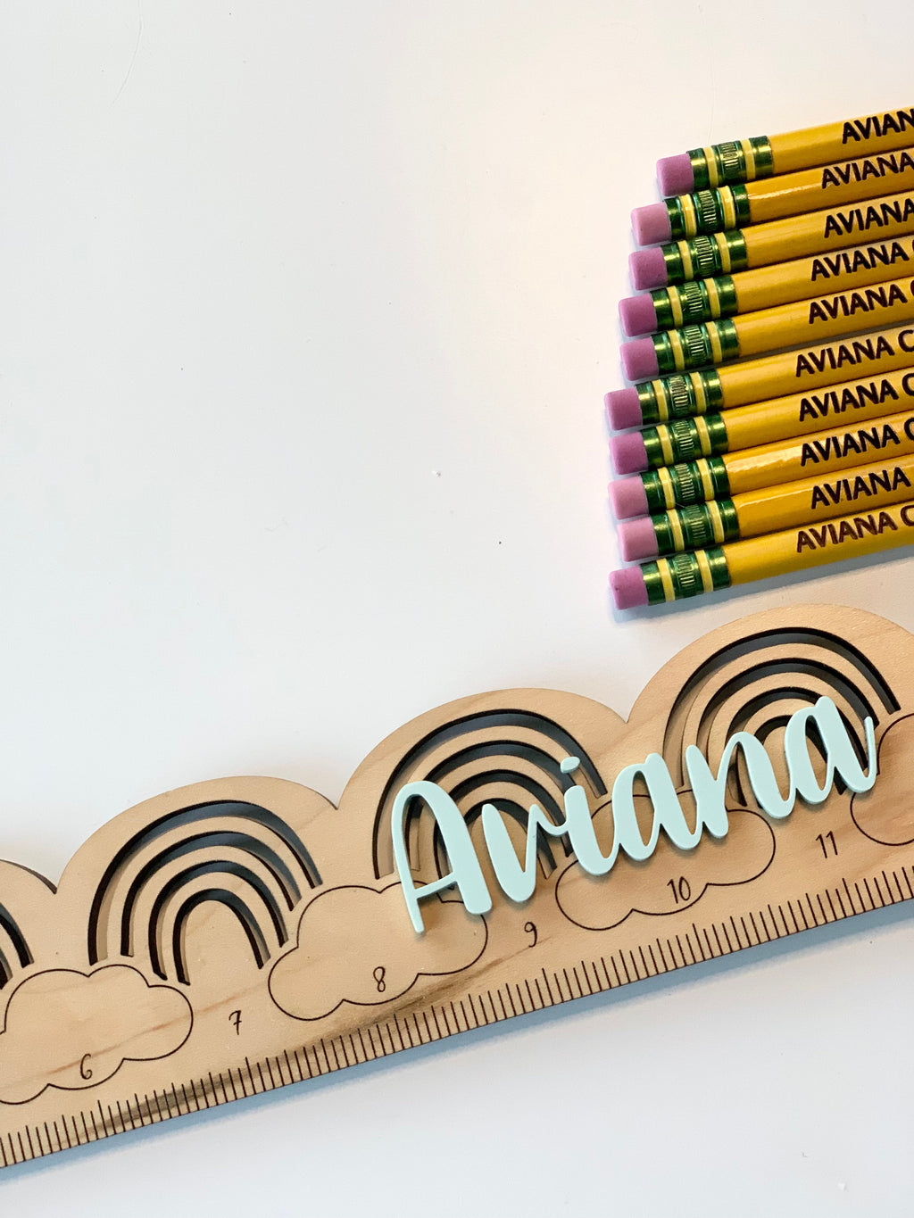 Set of personalized yellow pencils with pink erasers next to custom wooden ruler engraved with rainbows and name Ariana