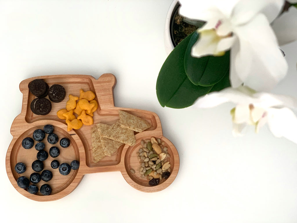 Natural wooden tractor-shaped sensory tray with sections holding blueberries, crackers, goldfish crackers, and seeds on white surface