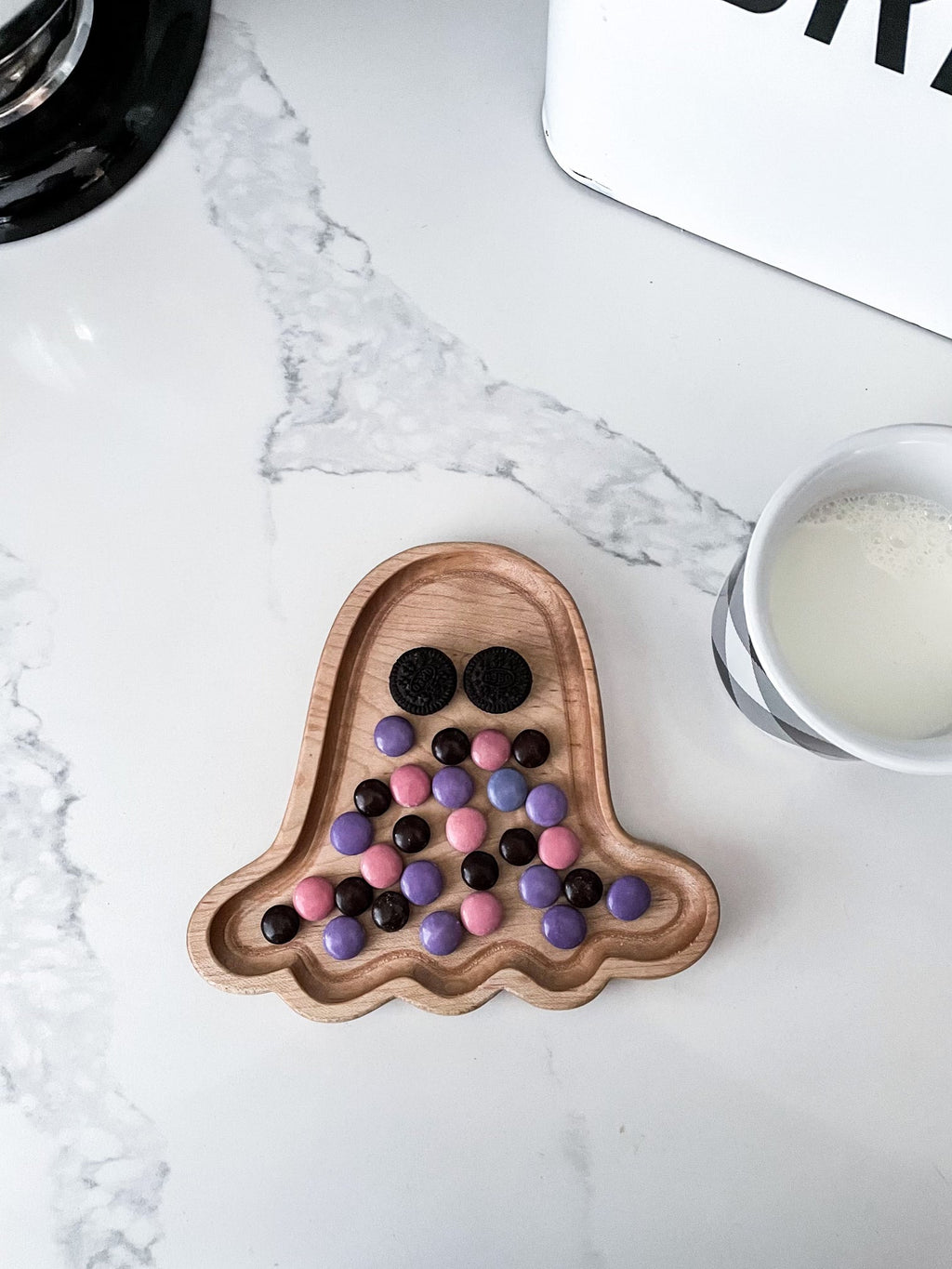 Small natural wood ghost-shaped sensory tray with colorful candy and black cookie eyes on white marble surface