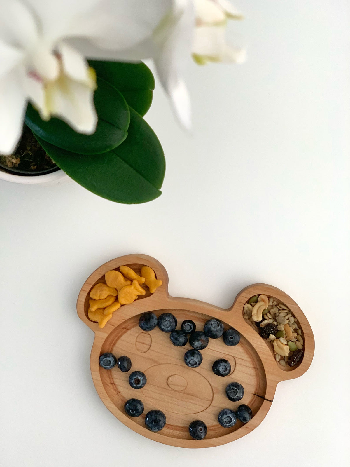 Natural wooden bear-shaped sensory tray with blueberries, goldfish crackers, and mixed nuts on white surface near plant leaves
