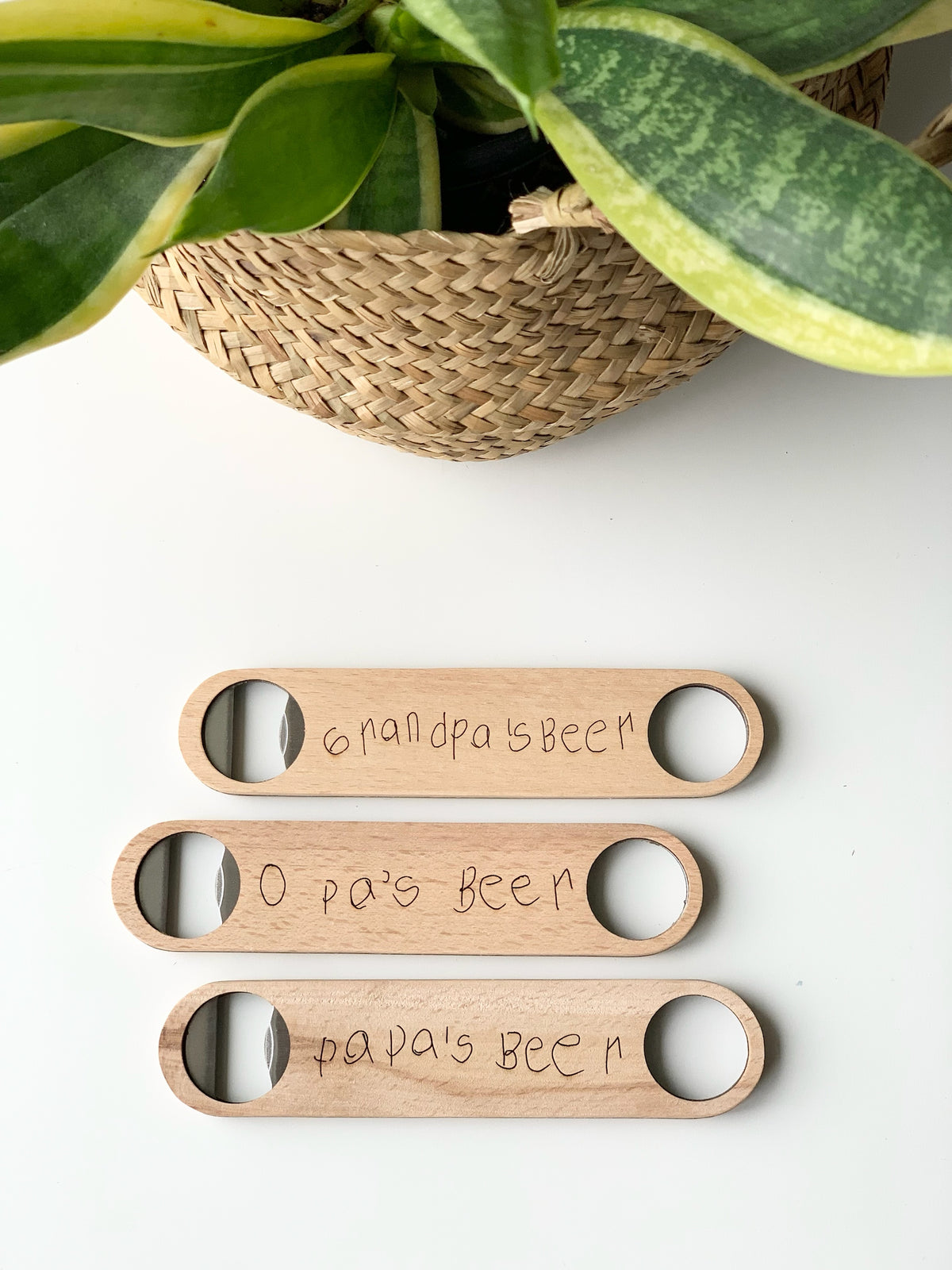 Three custom wooden bottle openers with child handwriting engravings on light wood laid on white surface near green plant