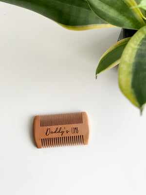 Small natural wooden beard comb with engraved "Daddy's Comb" text on white background near green plant leaves