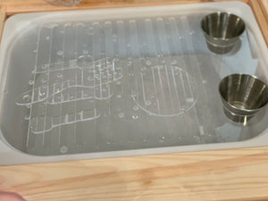 Plastic play kitchen sink insert with dish rack pattern and two metal play cups on wooden surface