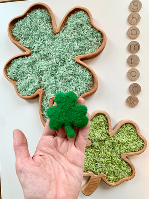 Hand holding a small green felt shamrock with two wooden shamrock trays filled with green rice and wooden letter tiles spelling shamrock