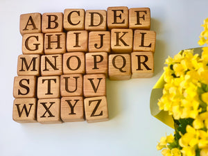 Natural 1.5-inch wooden alphabet blocks with engraved capital letters arranged in rows near yellow flowers on white surface