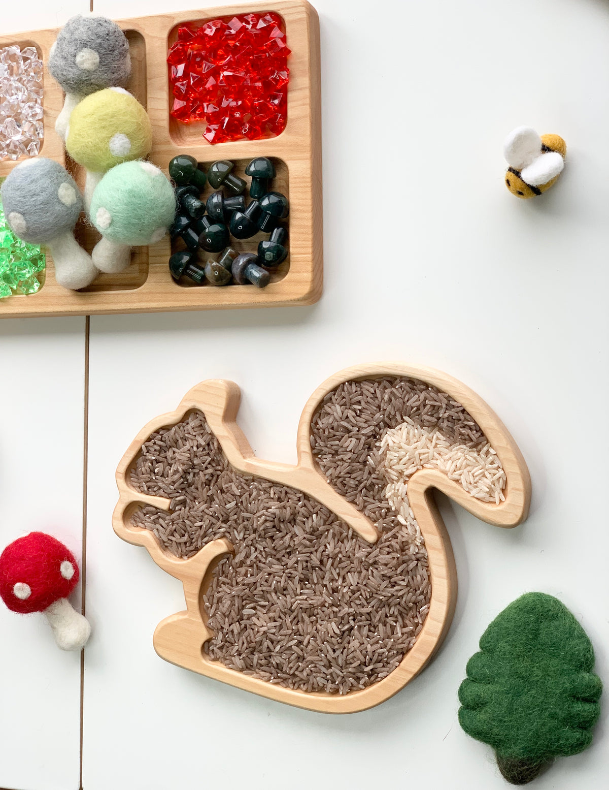 Wooden squirrel sensory tray filled with light and dark rice grains surrounded by felt mushrooms and bee toys on white table
