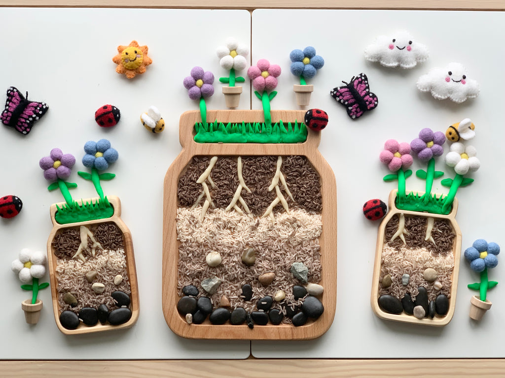 Large wooden sensory tray shaped like a jar with soil layers, roots, rocks, and colorful felt flowers on white background