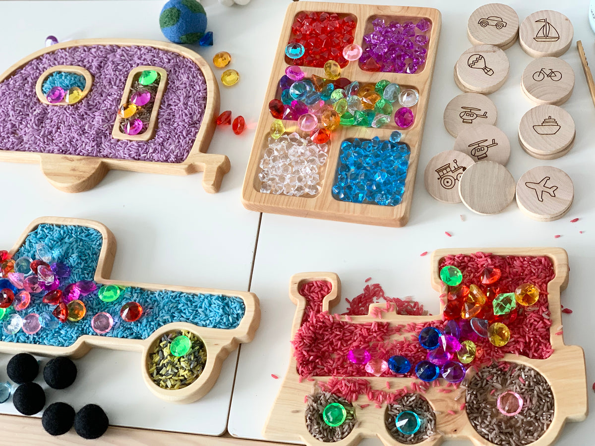 Wooden sensory trays shaped like a helicopter, truck, airplane, and train filled with colorful sensory materials and beads