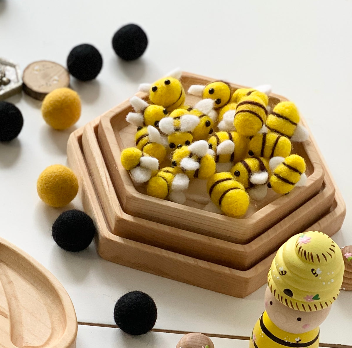 Natural wooden nesting hexagon plates stacked with yellow and black felt bees and scattered felt balls on white surface