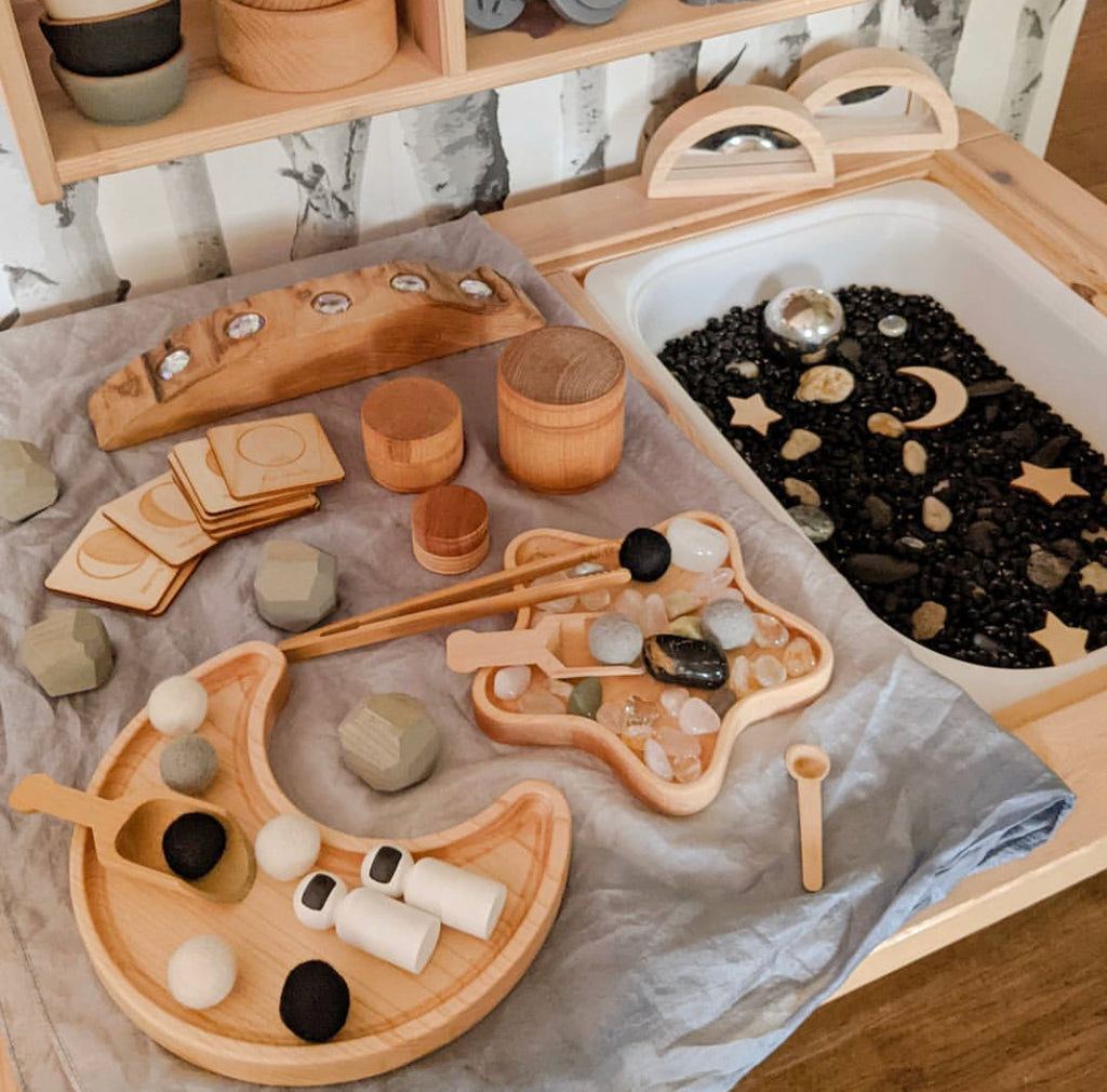 Moon-shaped wooden sensory tray with geometric and natural objects on linen cloth next to black sensory bin with celestial shapes