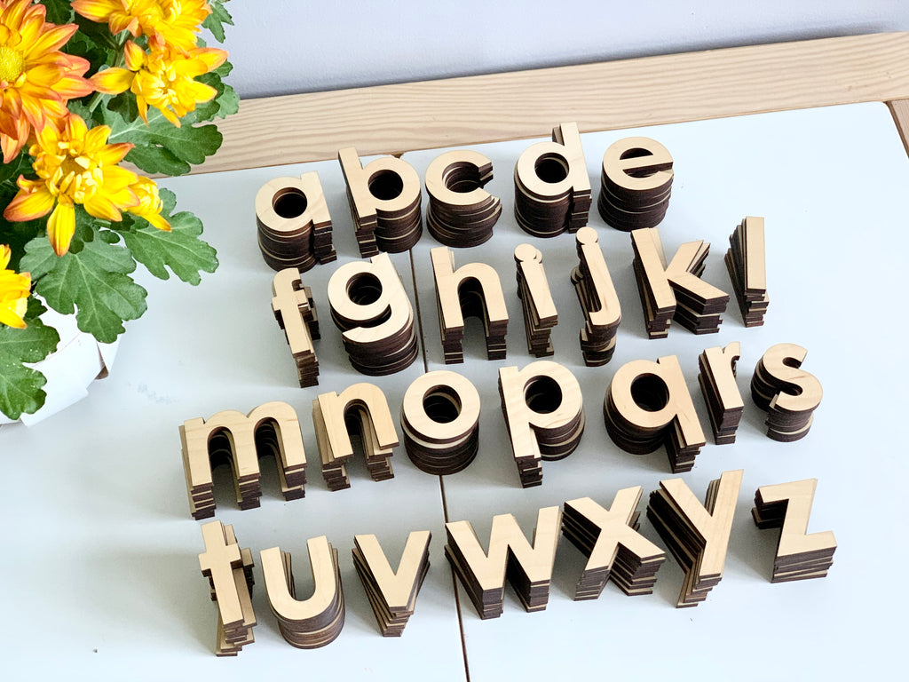Natural wooden lowercase alphabet letters stacked in neat rows on white surface with yellow flowers on side