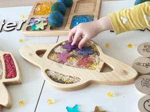 Natural wooden Saturn-shaped sensory tray filled with colored rice and star-shaped felt pieces being played with by child’s hand