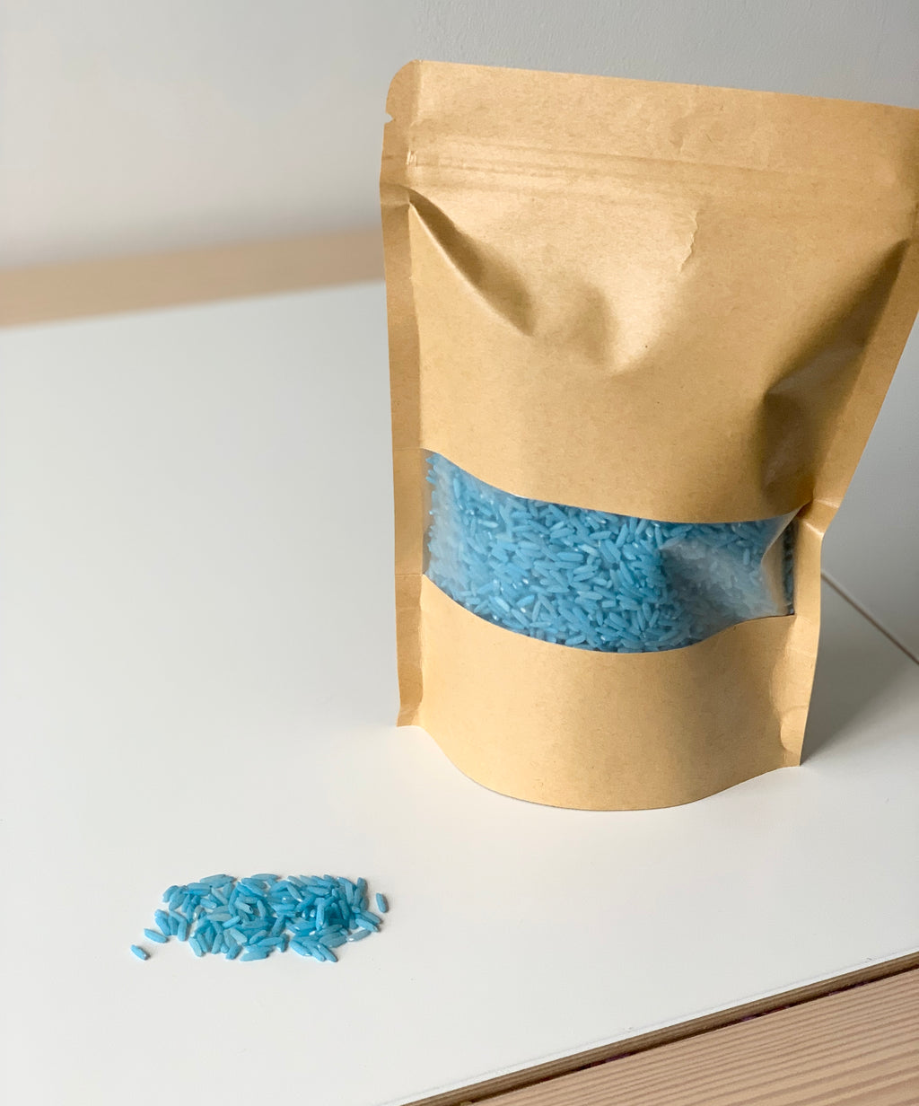 Brown paper pouch with clear window showing blue colored rice sensory filler on white surface with some spilled grains