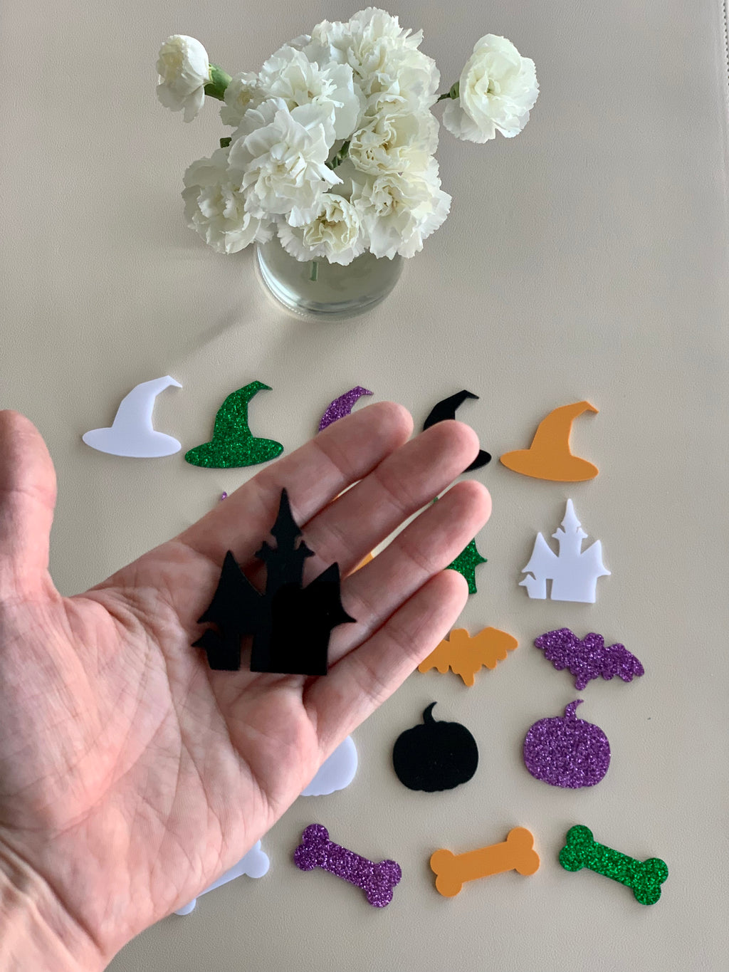 Hand holding black acrylic haunted house shape with various colorful Halloween acrylic shapes on beige surface and white flowers in glass vase above