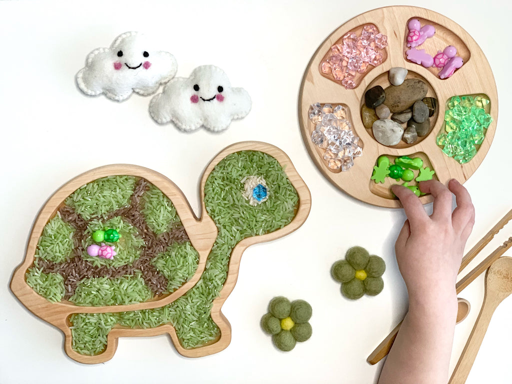Natural wooden turtle sensory tray filled with colored rice and a round tray with compartments holding gems and stones on white background