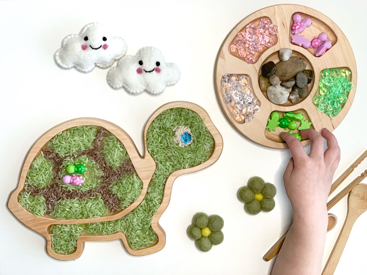 Natural wooden turtle sensory tray filled with colored rice and a round tray with compartments holding gems and stones on white background