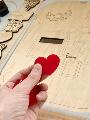 Hand holding red acrylic heart shape above wooden board with engraved heart and love letter shapes for sensory play