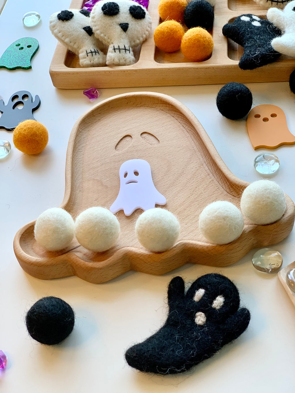 Natural wood ghost-shaped sensory tray with white felt balls and black and orange felt ghost and pumpkin decorations on white surface