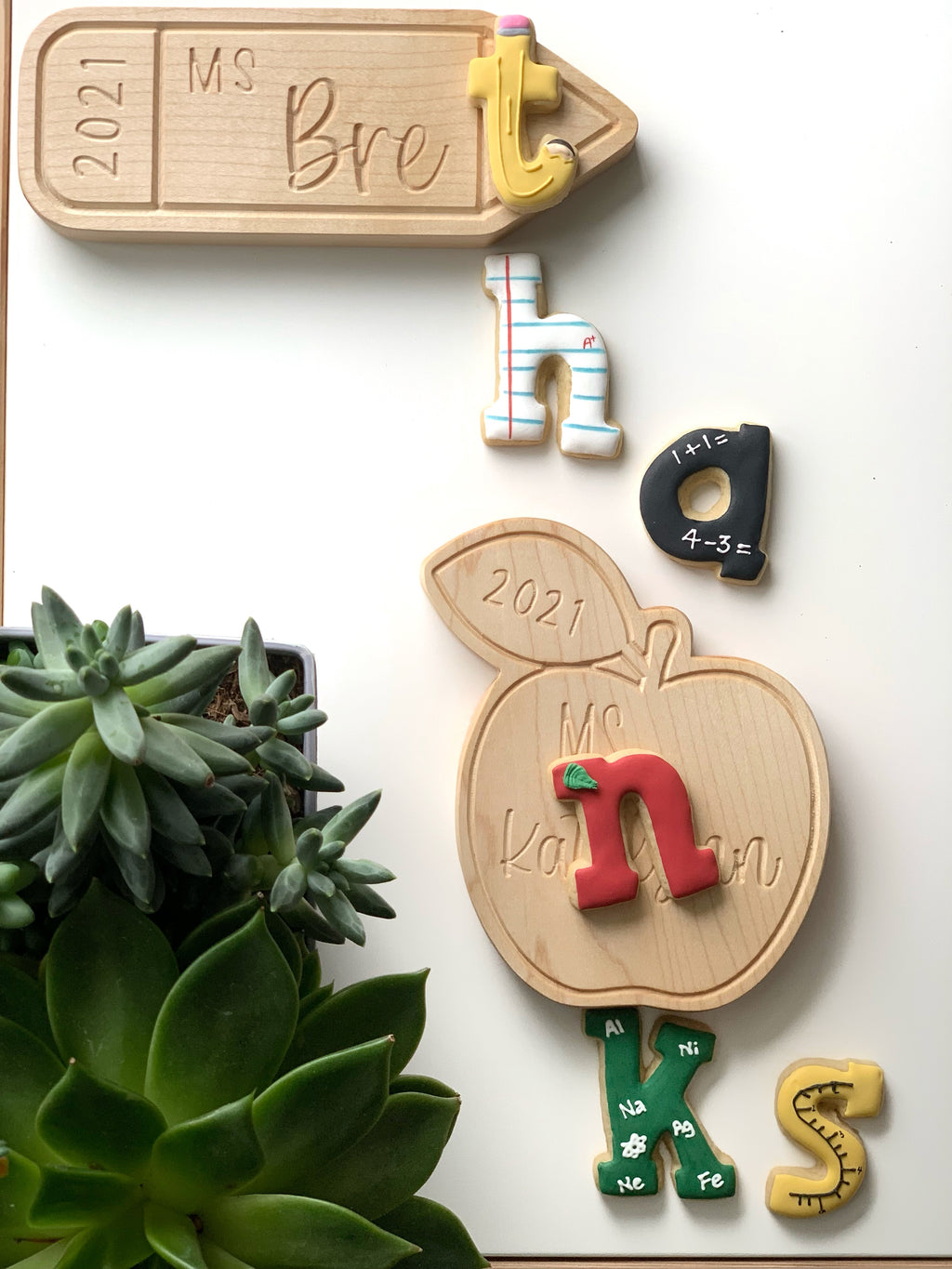 Personalized wooden teacher name plates in arrow and apple shapes with colorful alphabet cookies on white background with green succulent plants