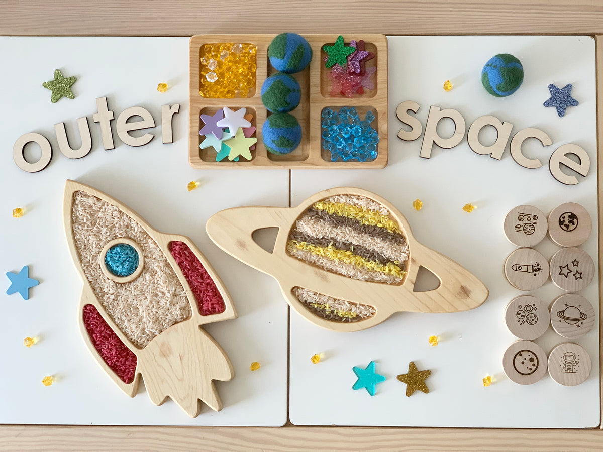 Wooden rocket ship and planet sensory trays with colorful fillers, wooden outer space letters, and space-themed stamps on table
