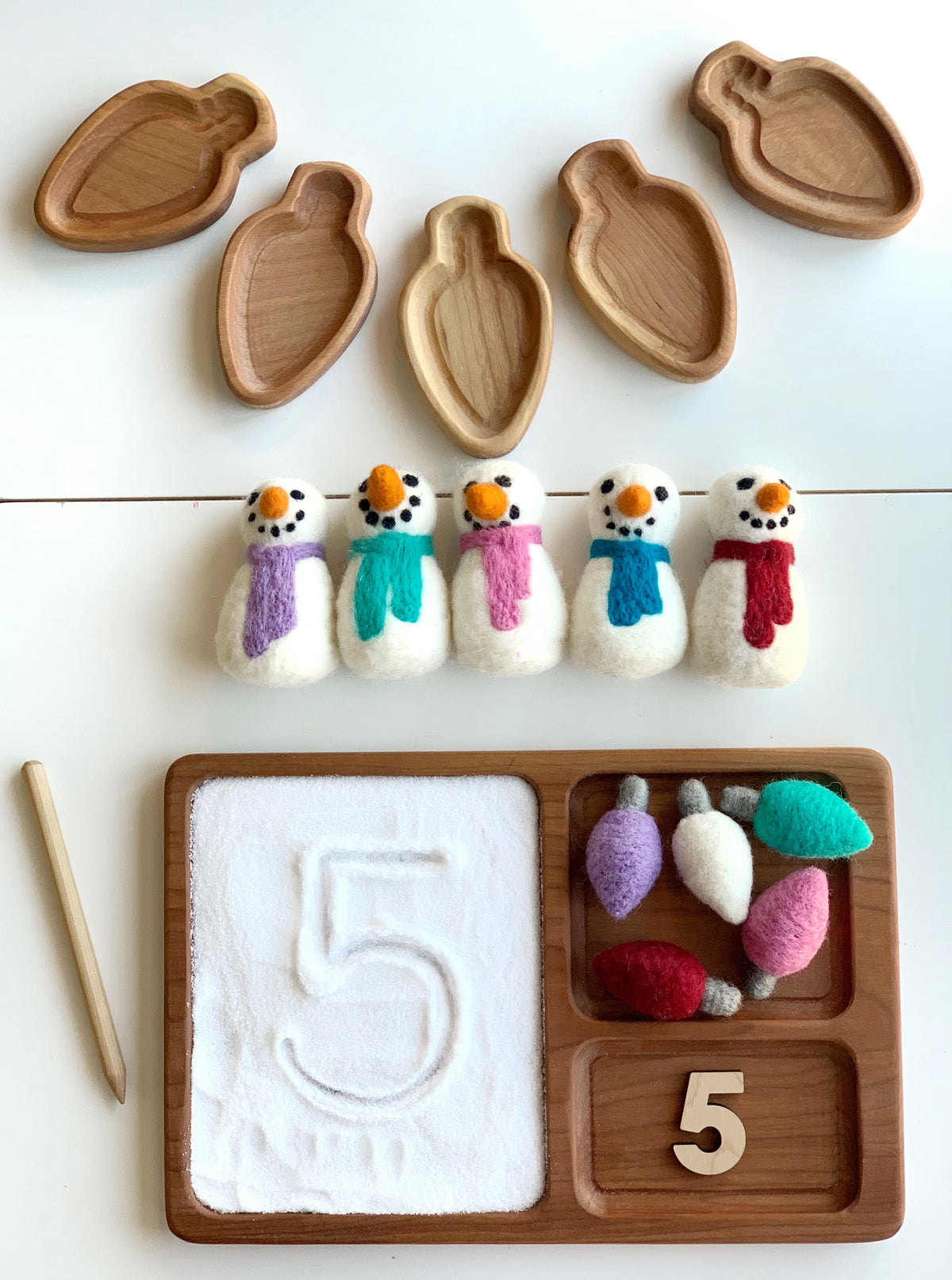 Natural wooden stylus with wooden tray containing white sand, felt snowmen, felt light bulbs, and number five cutout on white background