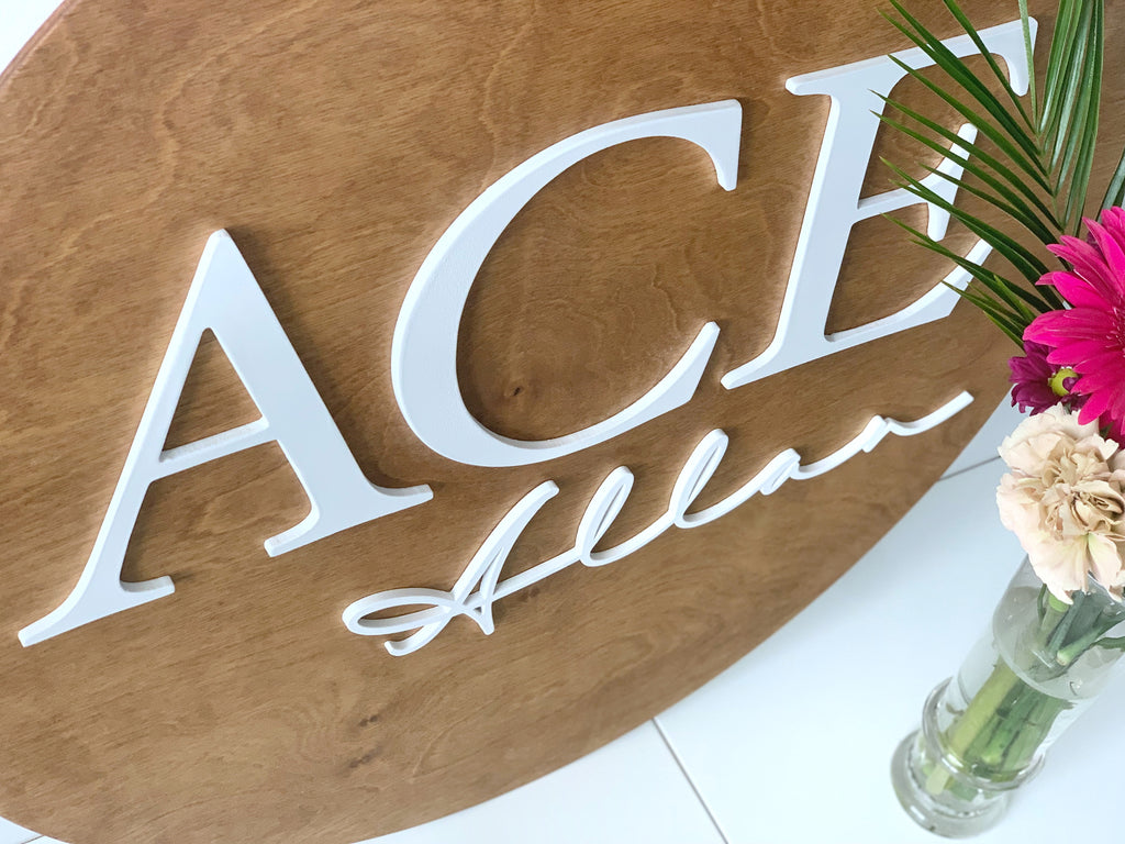 Large 3D round wooden name sign with white uppercase and cursive letters on natural wood background near flowers