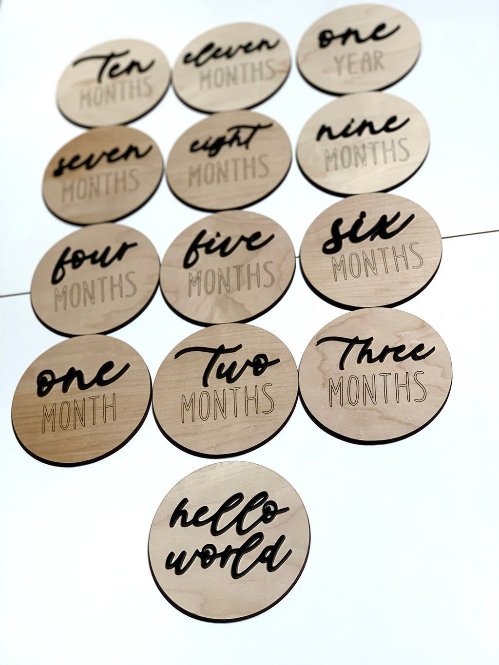 Natural wooden 3D milestone discs with black cursive and engraved letters displaying months and hello world text on white background