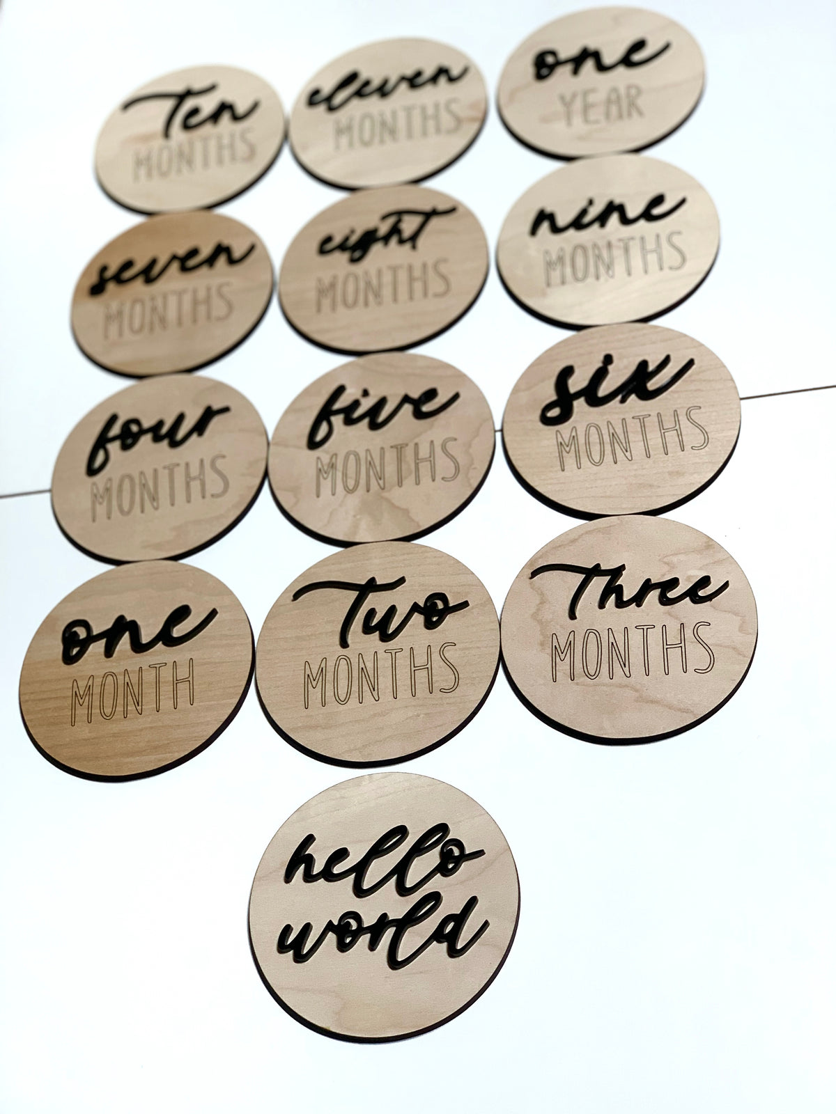 Natural wooden 3D milestone discs with black cursive and engraved letters displaying months and hello world text on white background