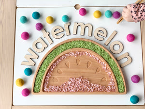Natural wooden watermelon sensory tray filled with green textured rice and pink beads on white table with colorful felt balls