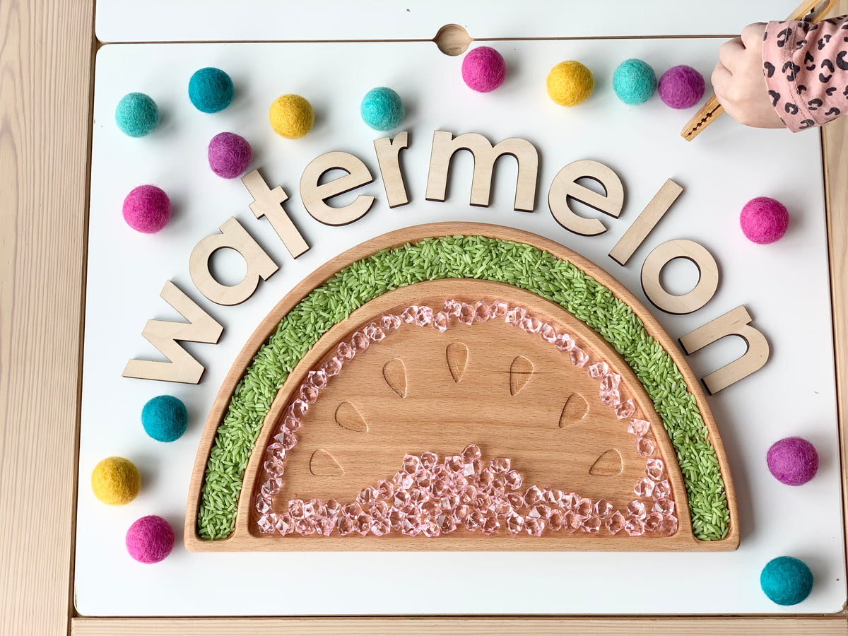 Natural wooden watermelon sensory tray filled with green textured rice and pink beads on white table with colorful felt balls
