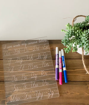 Acrylic cursive alphabet tracing board with engraved letters and three dry erase markers on wooden surface next to green plant