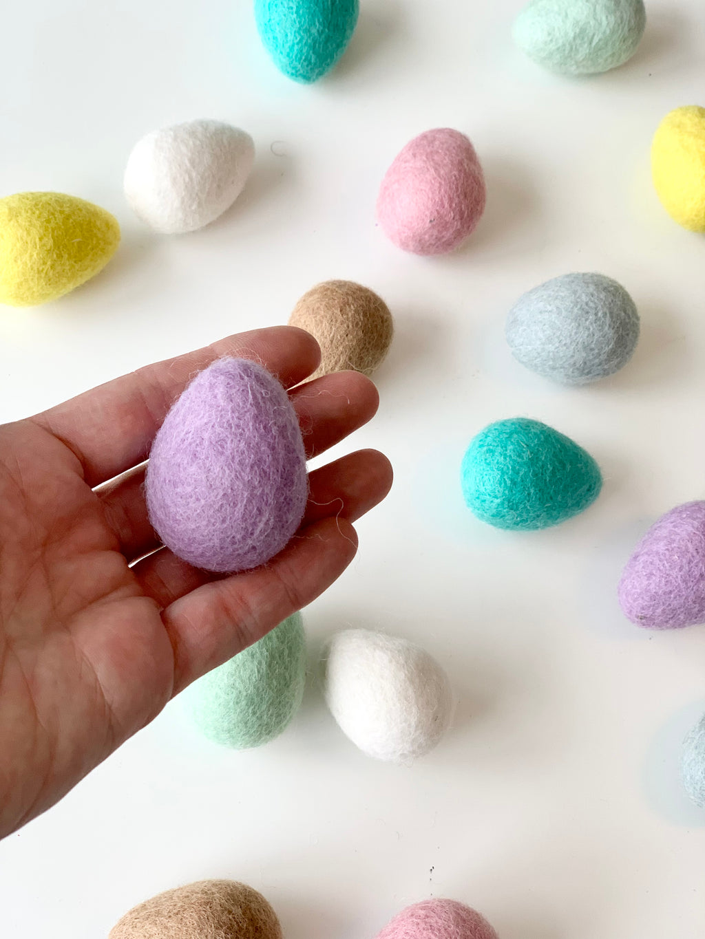 Hand holding single purple felt Easter egg with scattered pastel felt eggs on white background