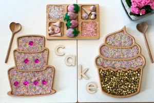 Wooden sensory cake and cupcake trays with colorful rice, gold bells, wooden letters spelling CAKE, and heart-shaped spoons on white table