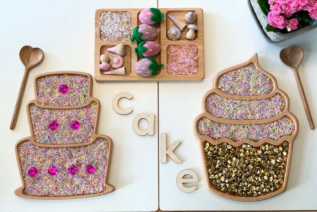 Wooden sensory cake and cupcake trays with colorful rice, gold bells, wooden letters spelling CAKE, and heart-shaped spoons on white table