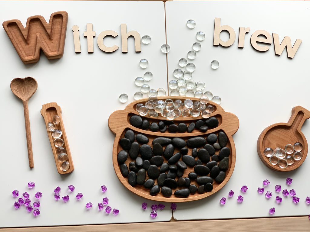 Natural wooden sensory tray shaped like a cauldron with black stones and clear beads bubbling on white background