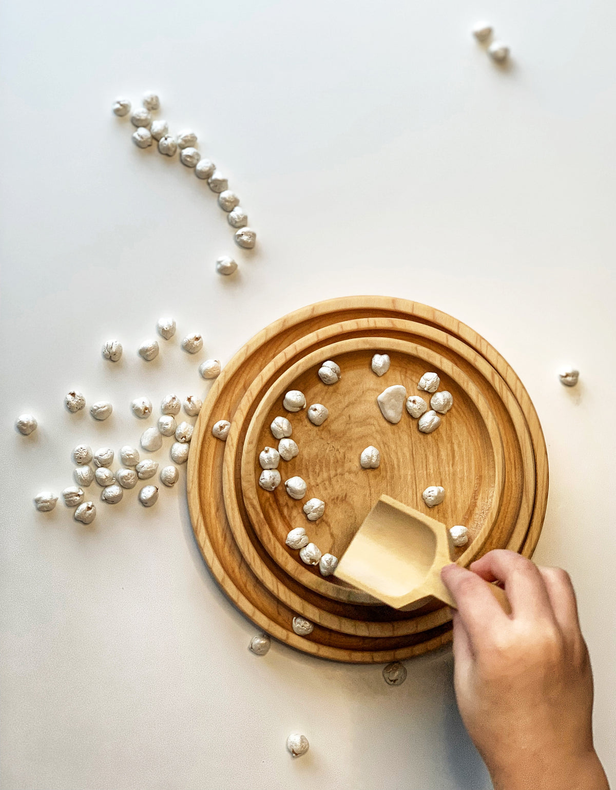 Three natural wooden nesting round plates stacked with small white textured beads and a hand holding a wooden scoop on white background
