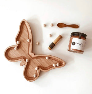 Natural wooden butterfly-shaped sensory tray with small wooden balls, jar of dough, small wooden spoon, and cork tube on white surface