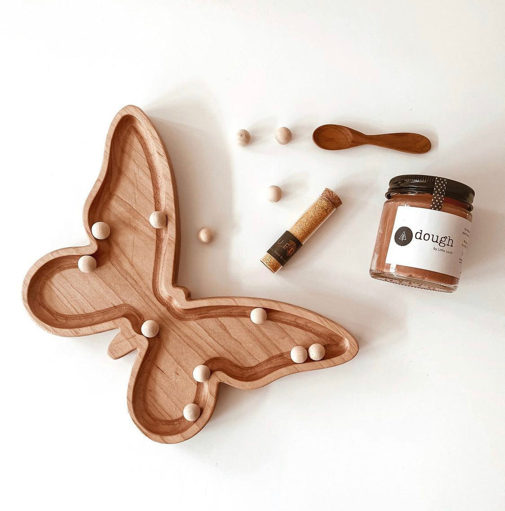 Natural wooden butterfly-shaped sensory tray with small wooden balls, jar of dough, small wooden spoon, and cork tube on white surface