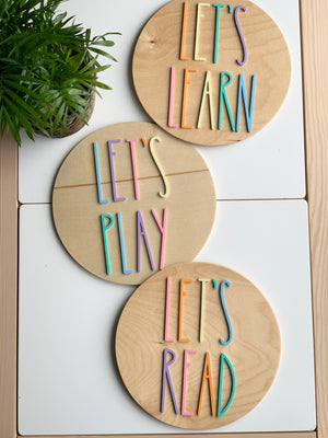 Set of three 10-inch round wooden signs with colorful pastel letters spelling Let's Play, Let's Read, and Let's Learn